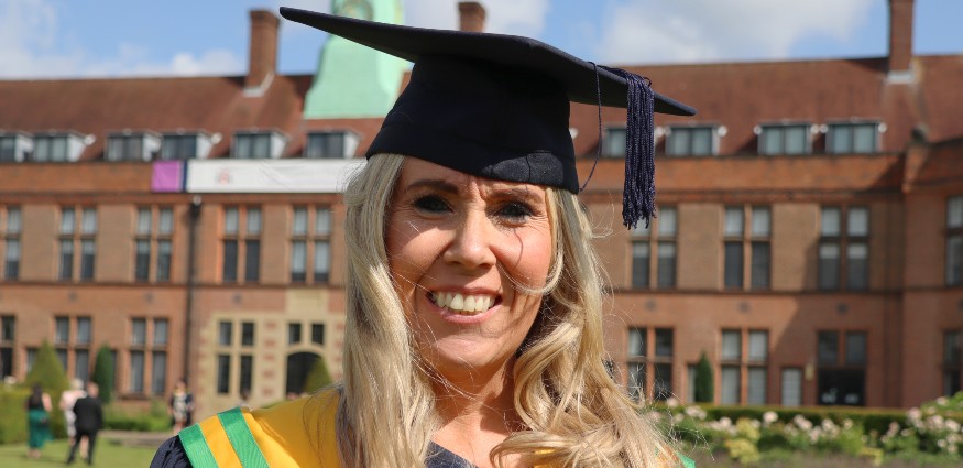 Emma Ellis wearing a graduation cap and gown, standing in front of the HCA Building at Hope Park.